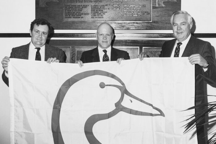 L’artiste commercial Ernie Wilson (au milieu), qui a créé le logo de CIC, tient un drapeau aux côtés de l’ancien vice-président exécutif de CIC, D.S. Morrison, et du président de CIC, Jack McKeag.